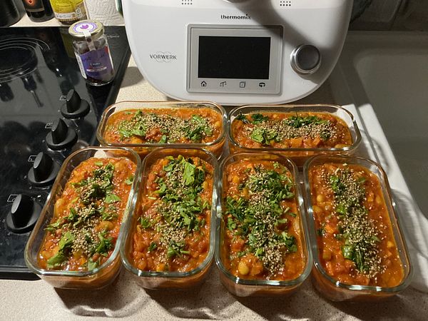 Six glass containers filled with a cooked bean dish garnished with herbs and seeds, placed on a kitchen counter beside a Thermomix appliance.