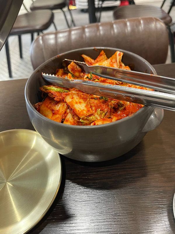 A bowl of kimchi is presented on a table with metal tongs resting on the edge.