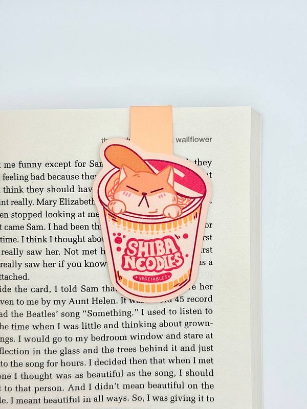A colorful magnetic bookmark featuring a cartoon cat in a noodle cup is placed on an open book page.