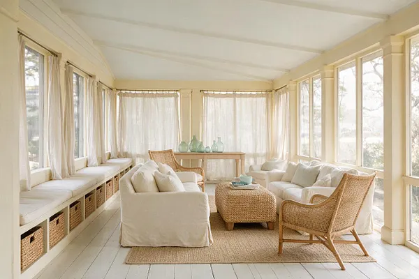 A bright enclosed front porch with white painted floors, sheer curtains, and wraparound windows. The space is furnished with slipcovered sofas, wicker chairs, a woven coffee table, and neutral cushions in a soft coastal palette.