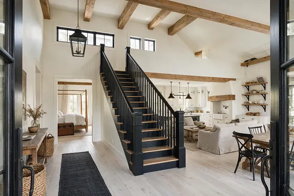 A bright farmhouse entryway with white walls, pale wood floors, and exposed ceiling beams. A black staircase rises in the center, with rustic lantern lighting, woven baskets, a dark runner rug, and an open view into a cozy living and dining area.