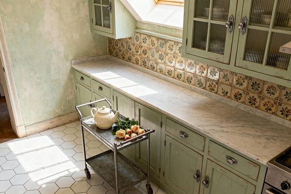A sunlit shabby chic kitchen with distressed mint-green cabinets and ribbed-glass uppers, topped by a long white marble countertop. A vintage-style floral ceramic tile backsplash, sage-washed plaster walls, and white hexagonal floor tiles set a soft palette, while a galvanized rolling cart with a cream teapot and roses sits below a skylight near a stainless range, creating a romantic cottage feel.