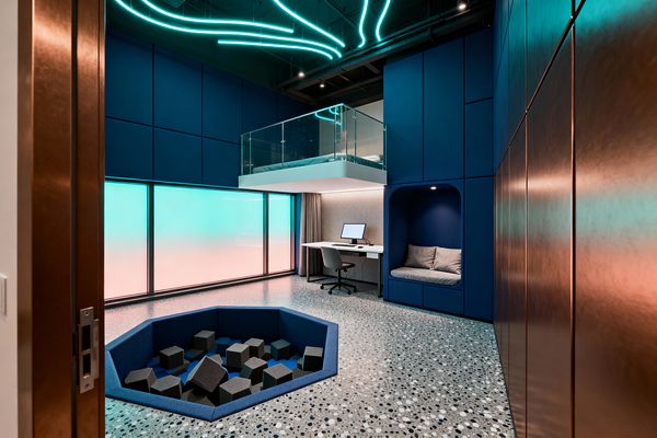 A futuristic kids’ room with deep blue wall panels features a sunken hexagonal play pit lined with padded sides and filled with dark foam cubes on a speckled terrazzo floor. A slim white desk with a computer sits beneath a floating mezzanine bed enclosed by a glass balustrade, next to a built-in alcove seat with gray cushions. Frosted backlit windows glow teal to peach, while curved turquoise LED strips sweep across the exposed black ceiling.