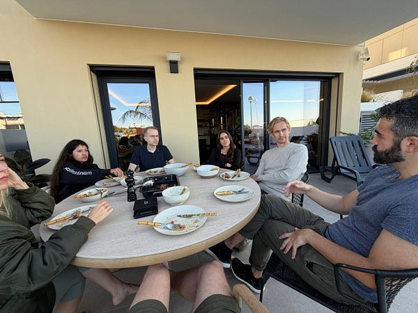 Group of six people sitting around a round outdoor table finishing a meal and having a discussion in a casual coworking meeting.