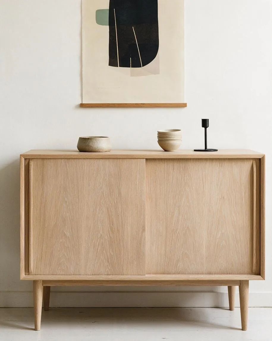 A light oak sideboard with slim tapered legs and sliding doors sits against a white wall. A simple abstract artwork hangs above it, and the top is styled with small ceramic vessels and a black candleholder.