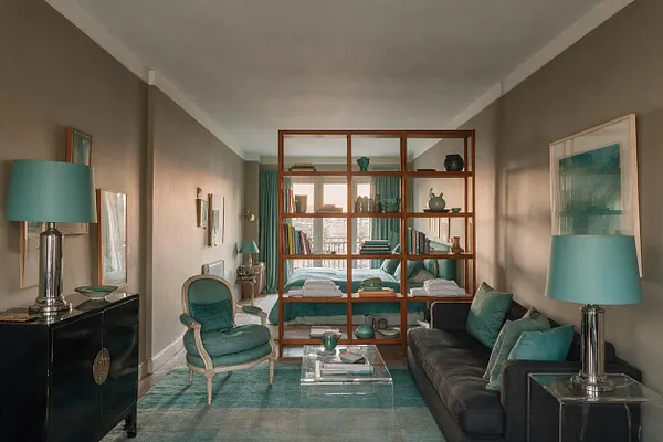 A narrow studio apartment combines sleeping and living areas with a wooden open shelving partition at the center. Teal accents appear in the lampshades, rug, pillows, and bedding, while a dark sofa, upholstered chair, and black cabinets frame the space under soft natural light.