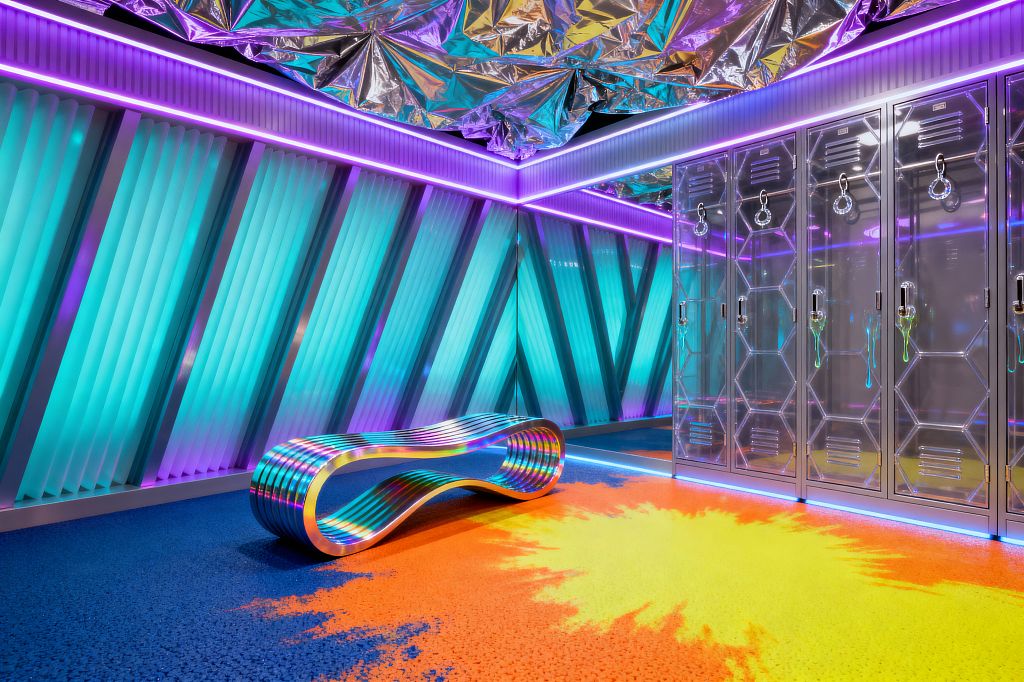 A neon-lit mudroom with ribbed translucent wall panels glowing in cyan and purple, outlined by continuous LED strips. The ceiling is covered in crinkled mirrored mylar, and a row of clear acrylic lockers with hexagon detailing and chrome ring pulls lines one side; at center sits an iridescent metal infinity-shaped bench on speckled flooring that gradients from electric blue to orange with a bright yellow burst, creating a futuristic, reflective atmosphere.