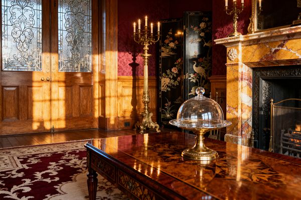A Victorian living room bathed in late-afternoon sun features carved oak paneling and double doors with ornate leaded glass. A gilded candelabrum glows beside a black lacquer Chinoiserie screen. The fireplace has a golden Siena marble mantel and a black cast-iron insert with a small fire. A burgundy-and-ivory patterned rug anchors an inlaid marquetry coffee table topped with a glass cloche on a brass stand.