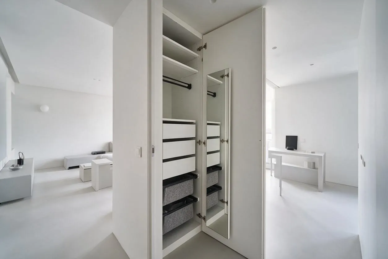 A minimalist built-in closet with a tall hinged mirrored door, open white shelving, a black hanging rod, and stacked white drawers with dark trim. The closet is set within a bright white room with clean lines and sparse furnishings, creating a calm, airy atmosphere.