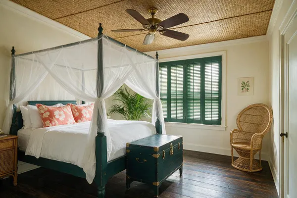 A tropical-style basement bedroom features a teal four-poster canopy bed draped with sheer white mosquito netting, white bedding, and coral floral pillows. The room has woven-texture ceiling material, dark wood floors, green shutters, a wicker chair, and a dark trunk at the foot of the bed.