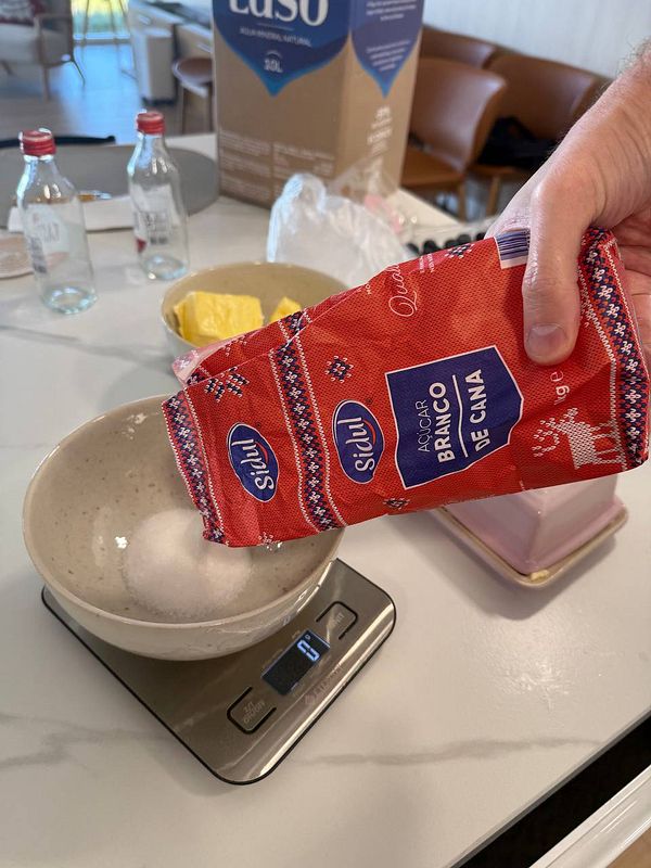 A person is pouring sugar from a red bag into a bowl on a digital scale while preparing to make butter cake.
