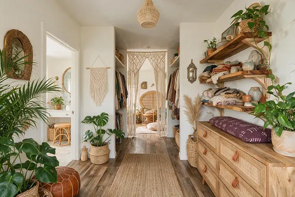 A bright bohemian closet with white walls, wood flooring, open wooden shelves, and hanging clothes arranged on both sides of a narrow walkway. The space includes a woven pendant light, macramé curtain and wall hanging, rattan chair, woven baskets, potted greenery, and a rustic wooden dresser, creating a warm, natural, layered look.