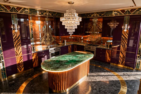 Opulent Art Deco kitchen with a U-shaped layout featuring deep aubergine lacquered cabinets inset with zebrawood panels, brass trims, and stainless appliances. A tiered crystal chandelier hangs over an oval island with an emerald green onyx top and fluted wood base, while mirrored and copper geometric backsplashes, stained-glass frieze, warm under-cabinet lighting, and a black marble floor with circular brass inlay complete the scene.