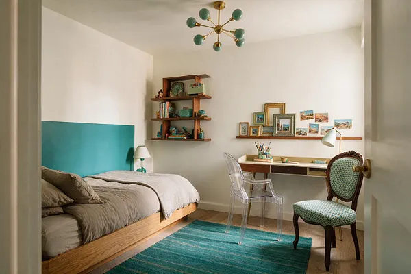 A bright kids' room with white walls, a low wooden bed with gray bedding and a teal headboard panel, a turquoise area rug, and a small study desk along the right wall. Floating shelves display books, art, and framed pictures, while a transparent chair and a vintage-style upholstered chair add eclectic character.