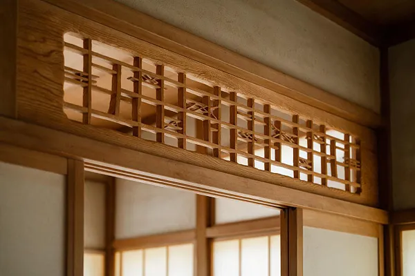 A close-up of a Japanese-style interior showing a carved wooden transom with a geometric lattice pattern above a sliding doorway. The warm wood trim contrasts with soft neutral wall panels and frosted light behind the opening, creating a calm Zen atmosphere.