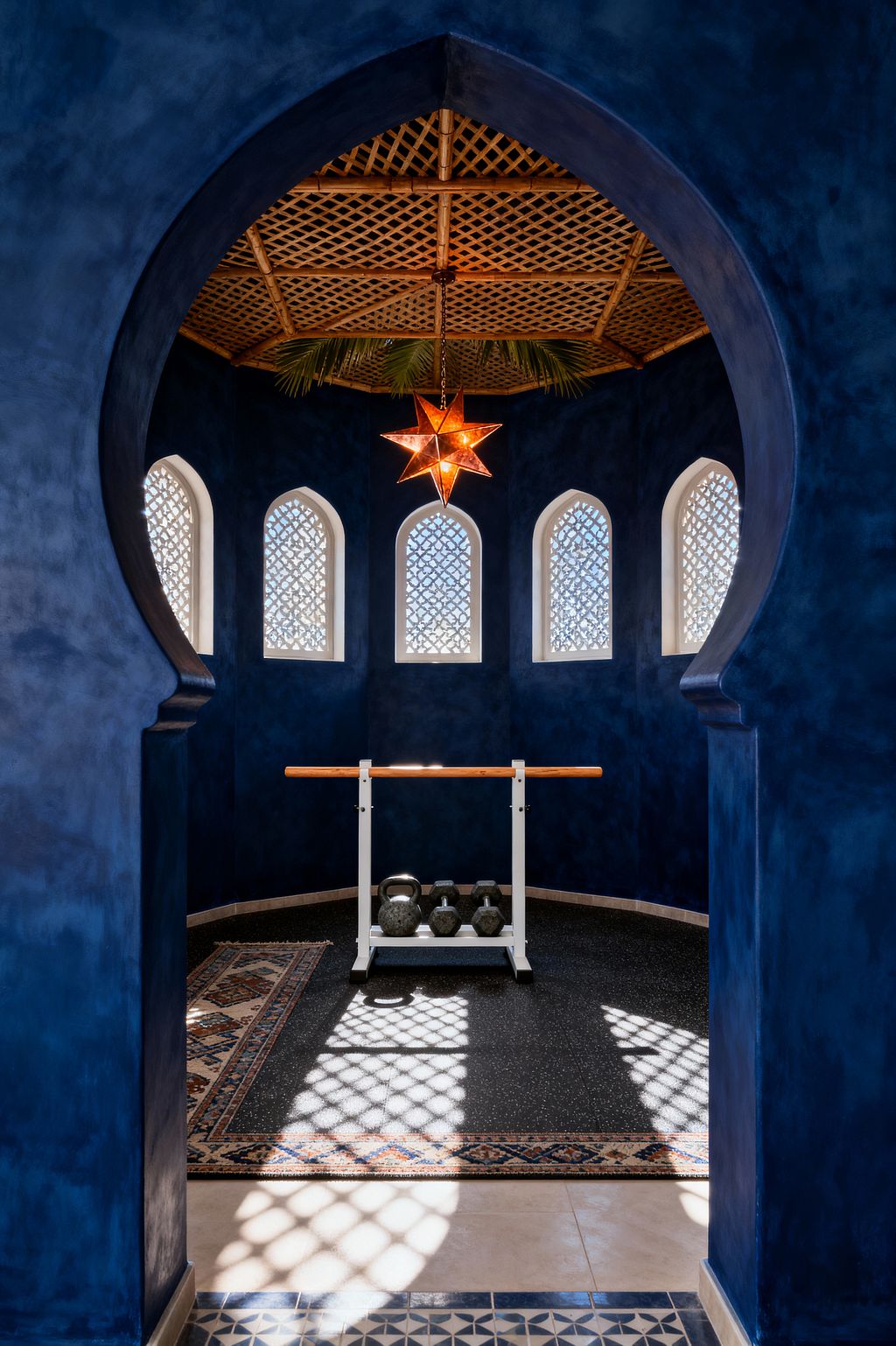 A view through a keyhole-shaped arch shows a compact home gym with deep cobalt plaster walls and an octagonal layout. Five arched windows with white lattice screens cast patterned light onto black speckled rubber flooring edged by a patterned rug. A wooden ballet-style barre on a white metal stand faces a row of kettlebells. Above, a woven rattan ceiling with palm fronds centers a glowing brass-and-amber Moroccan star pendant.