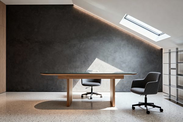 A contemporary home office features a glass-top desk on a light wood base centered on a pale terrazzo floor. Two gray upholstered swivel chairs with black casters sit before a charcoal microcement wall. A sloped ceiling with a skylight and concealed linear LED washes the wall and floor with daylight. To the right stands open stainless-steel shelving against a white partition, while vertical wood slats on the left complete the clean, minimalist space.