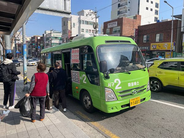 A small green bus is parked at a city street stop with passengers boarding.