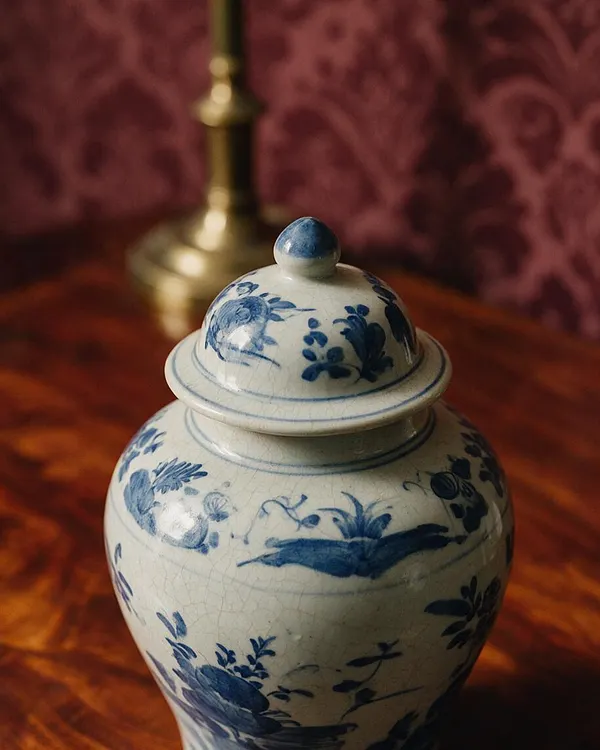 A traditional blue and white ceramic ginger jar with a domed lid sits on a polished wooden surface. The jar features hand-painted floral motifs, with a warm brass lamp base and burgundy patterned wallpaper softly blurred in the background.
