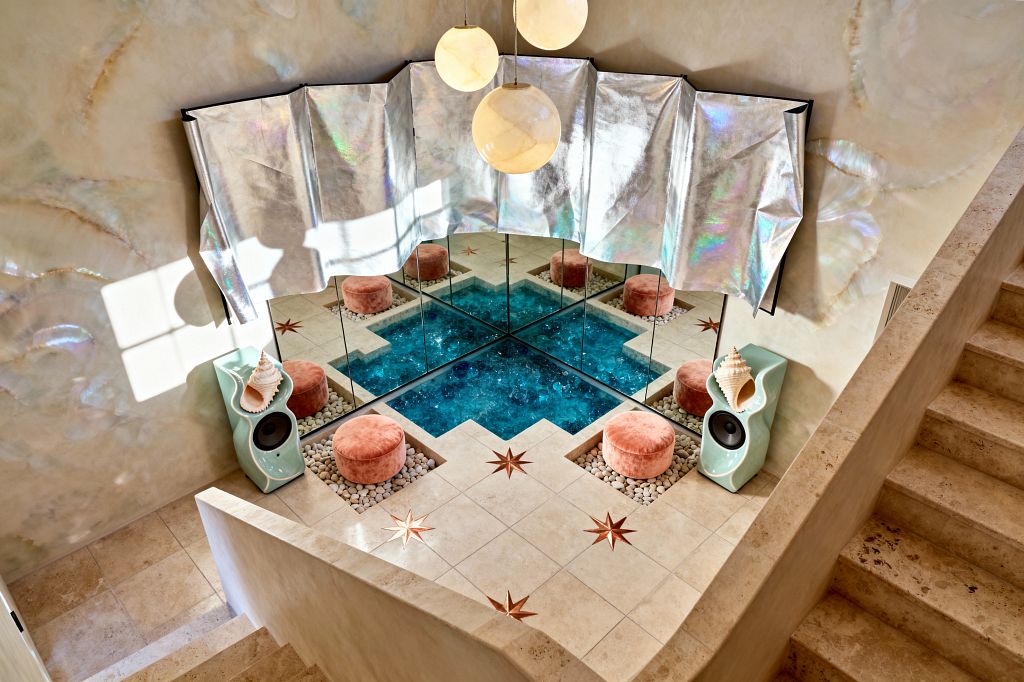 Media room with a sunken square lined by mirrors that reflect a blue glass pool at the center. A sand-toned travertine floor features star inlays and recessed pebble beds holding round blush velvet poufs. On each side, mint green sculptural speakers sit with large conch shells on top. Three creamy globe pendants hang above, while an iridescent silver drape and pearlescent plaster walls glow softly. Stone steps descend into the intimate space.