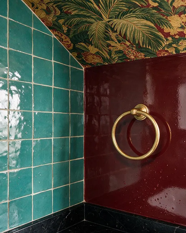 A close-up of a maximalist bathroom corner with glossy teal square wall tiles, a deep burgundy lacquered surface, and a brass towel ring. The ceiling is covered in dense tropical floral wallpaper, and a black stone ledge anchors the bottom of the composition.