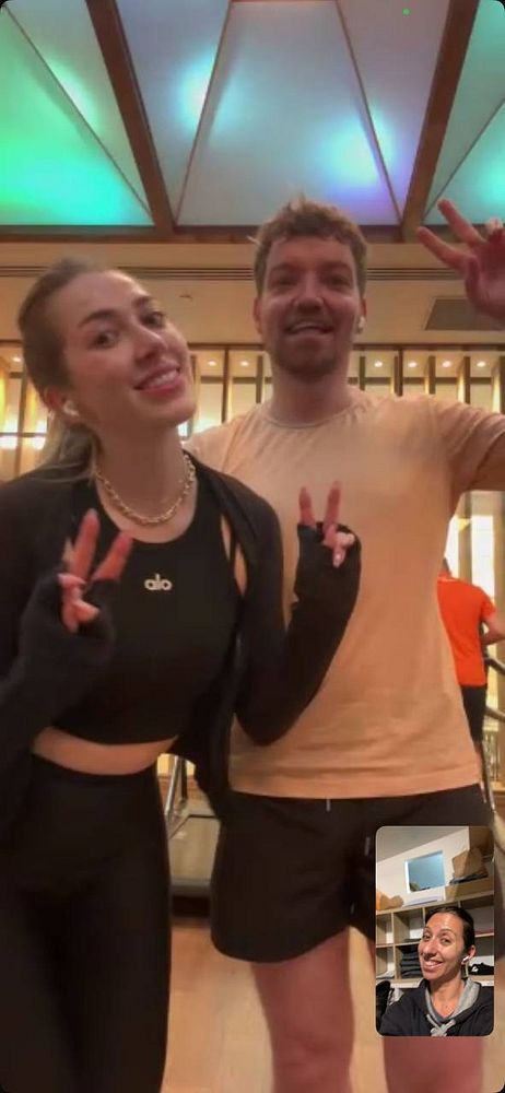 Two individuals pose for a cheerful photo in a gym setting while video chatting with a third person.