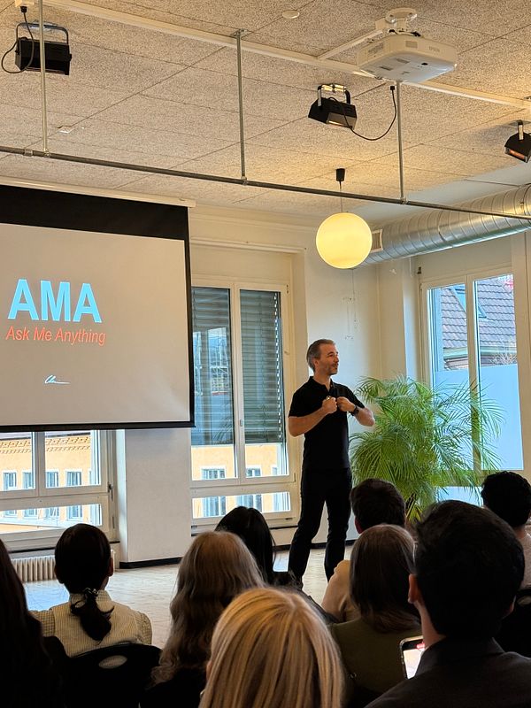 A speaker is presenting during an AMA session in a well-lit room with an audience.