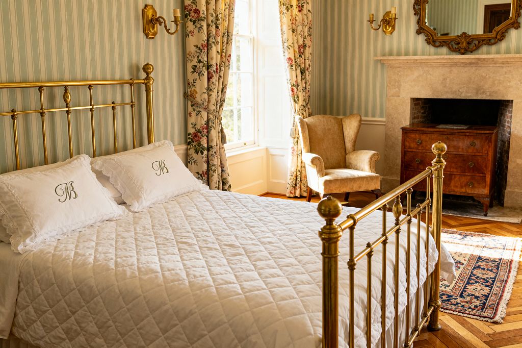 A traditional bedroom features a polished brass bed with white quilted bedding and monogrammed pillows beside a tall sash window. Pale aqua striped wallpaper, floral chintz curtains with tiebacks, brass candle sconces, a beige wingback chair, a limestone fireplace topped by a gilt mirror, a mahogany chest, a Persian-style rug, and oak herringbone floors are illuminated by warm daylight.
