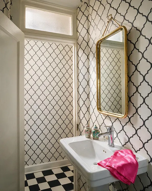 A small Hollywood Regency powder room with cream walls covered in bold black quatrefoil wallpaper, a gold-framed wall mirror, and a white pedestal sink. The floor is black-and-white checkerboard tile, and bright pink fabric adds a vivid accent near the chrome faucet.