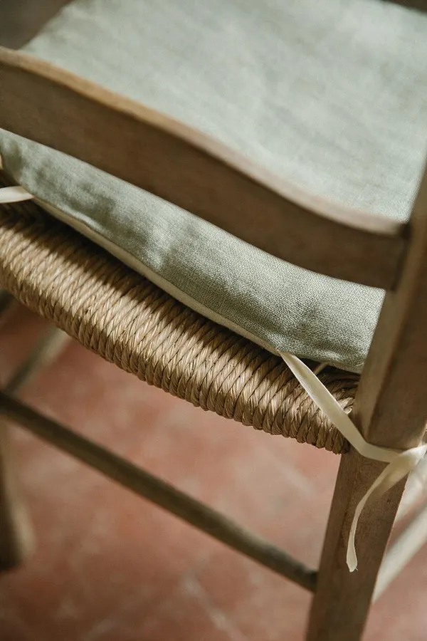 A close-up of a French Country chair showing a pale linen seat cushion tied to a natural wood frame with cream ribbon. The woven rush seat and terracotta floor create a warm, rustic, handcrafted look.