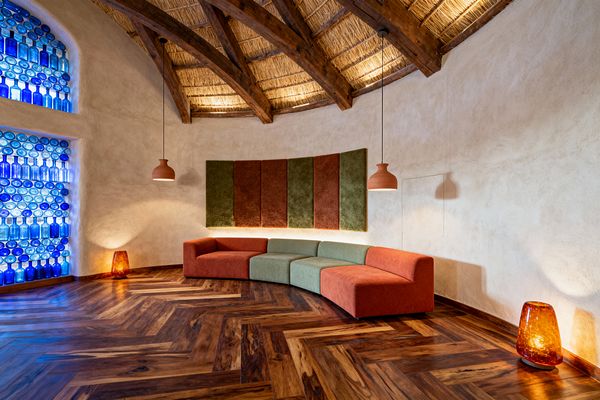 A Southwestern-inspired media room features a curved modular sofa in terracotta and sage set against cream plaster walls beneath a thatched ceiling with exposed timber beams. A herringbone hardwood floor and terracotta pendant lights warm the space, while an arched wall of cobalt blue glass bottles filters daylight and vertical fabric acoustic panels echo the earth-toned palette, creating an intimate, minimalist setting.
