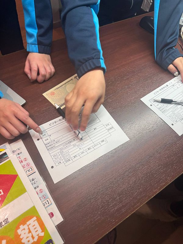 The image captures a scene at a reception desk where individuals are filling out forms for a gym day pass in Japan.