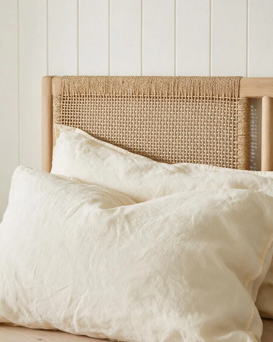 A coastal-style bed features a light wood frame and a woven cane headboard panel behind layered cream-colored linen pillows. The background is a simple white paneled wall, and the overall look is airy, natural, and relaxed.