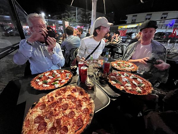 A casual group photo of four men sitting and standing around a table with several pizza boxes and drinks in a well-lit indoor setting.