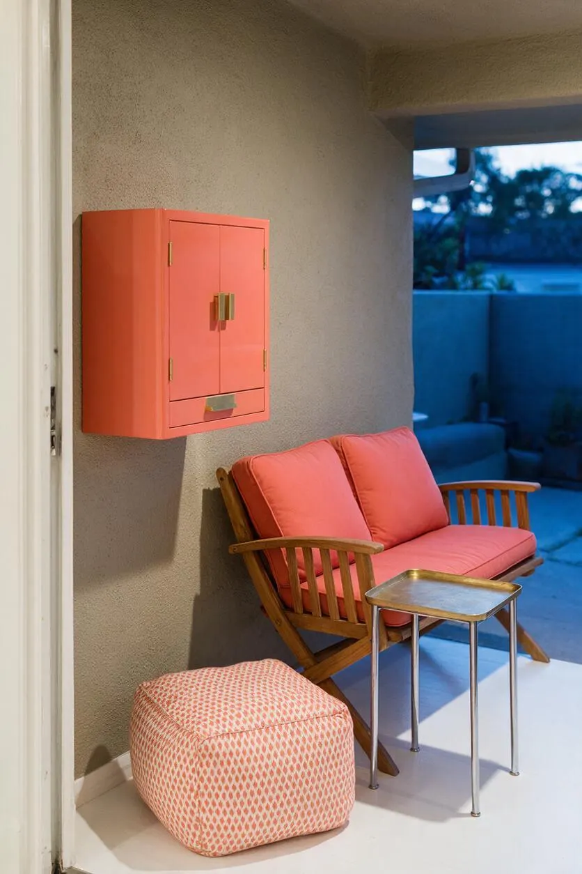 A compact eclectic patio features a coral wall cabinet mounted on a textured stucco wall, a wooden slatted loveseat with coral cushions, a small brass-topped side table, and a patterned pink ottoman on a pale floor. The space opens to a blue-toned outdoor view, creating a colorful, relaxed sitting area.