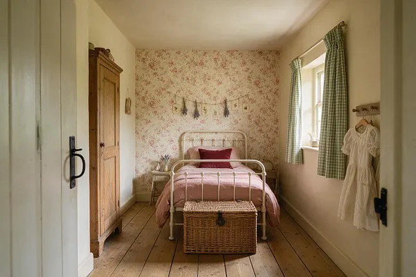 A cozy kids' room with floral wallpaper, a white metal bed dressed in pink bedding, and a woven storage chest at the foot of the bed. Green gingham curtains frame a window, while a wooden wardrobe and a small hanging dress add to the cottage-inspired charm.