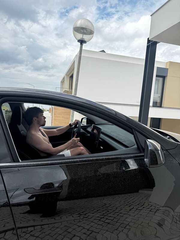 A shirtless man is sitting in the driver's seat of a black car parked in a residential area.
