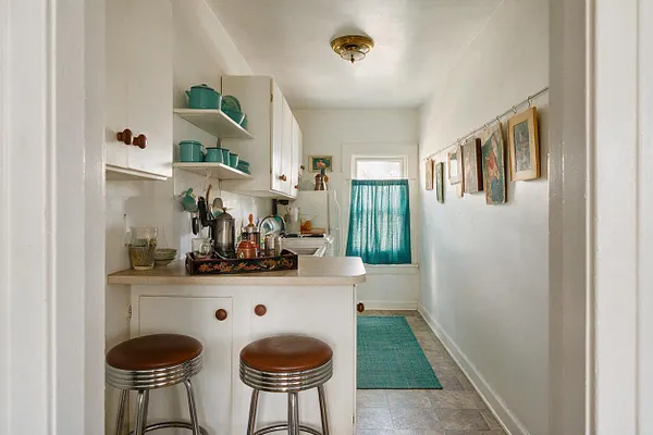 A narrow eclectic kitchen with white cabinets, open shelves holding teal cookware, and a small counter with two brown-and-chrome stools. Framed artwork lines the right wall, and a teal curtain and rug add color at the far end of the room.