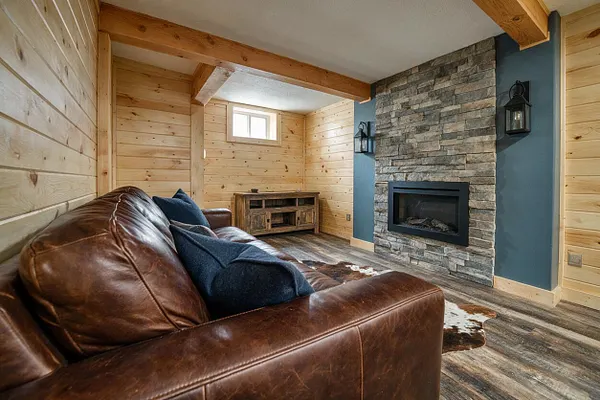 A cozy rustic basement lounge with knotty pine plank walls, exposed wood ceiling beams, and a stacked stone fireplace with a black insert on a blue accent wall. A brown leather sofa with dark blue pillows sits in the foreground, while a small wood media cabinet and wood-look floor complete the warm, cabin-inspired space.