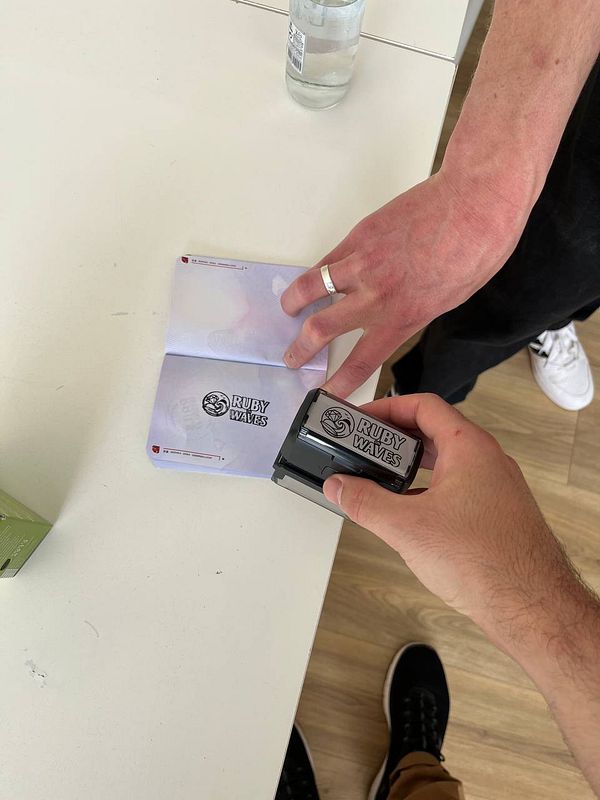 Close-up photo of a hand holding a rubber stamp featuring the Ruby on Waves logo with a clear plastic handle.