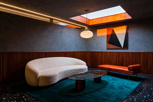 A modern living room with charcoal plaster walls above wood slat wainscoting, furnished with a curved ivory bouclé sofa, an oval glass coffee table on wood bases, and a burnt-orange leather daybed on thin metal legs. A deep teal rug lies on dark terrazzo flooring, while a white globe pendant, a terracotta-lined skylight, a narrow clerestory window, and a geometric artwork complete the warm, minimalist composition.
