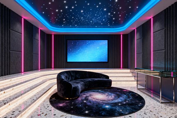 A futuristic media room features a fiber‑optic star ceiling bordered by blue cove lighting, charcoal acoustic wall panels, and vertical neon‑pink light strips. Curved terrazzo steps with integrated warm LEDs surround a crescent midnight‑blue velvet sofa on a round galaxy‑print rug facing a large wall‑mounted screen; a slim glass‑topped chrome console sits along the right wall, with reflective terrazzo flooring completing the immersive look.