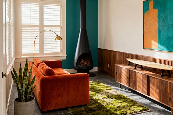 A mid-century modern living room with a rust-orange corduroy sofa facing a black cone fireplace set against a teal accent wall. Sunlight from white plantation-shuttered windows highlights walnut slat wainscoting, a low walnut credenza displaying a small surfboard, a brass arc floor lamp, a moss-green shag rug on dark slate tile, and a snake plant in a concrete pot.