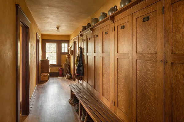 A long Craftsman-style mudroom with a wall of built-in oak cabinets on the right, featuring paneled doors, dark hardware, and a slatted wood bench. The space has warm golden walls, a wood floor, a small window at the far end, and practical hooks, baskets, and pottery accents.