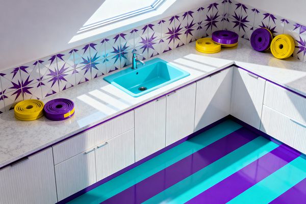 A pop art–style laundry room with white ribbed cabinets and pale speckled quartz counters features a bright turquoise drop-in sink with a chrome faucet. The backsplash is white ceramic tile printed with purple starbursts, purple trim outlines the cabinetry, and a high-gloss floor alternates broad teal and purple stripes; a skylight casts diagonal shadows across the counter, and coiled yellow and purple accessories rest near the corner.
