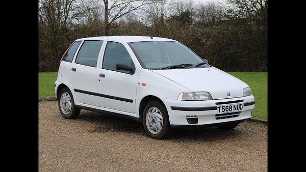 A white hatchback car is parked on a gravel surface with greenery in the background.