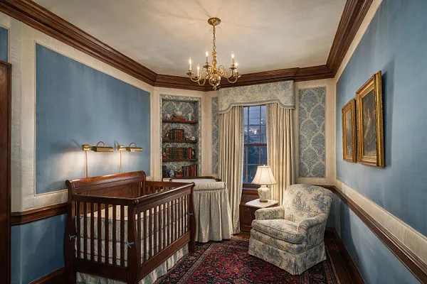 A traditional nursery with blue walls, cream trim, and dark wood crown molding. The room includes a wooden crib, a patterned upholstered armchair, a small lamp by the window, built-in bookshelves, and a classic chandelier overhead.