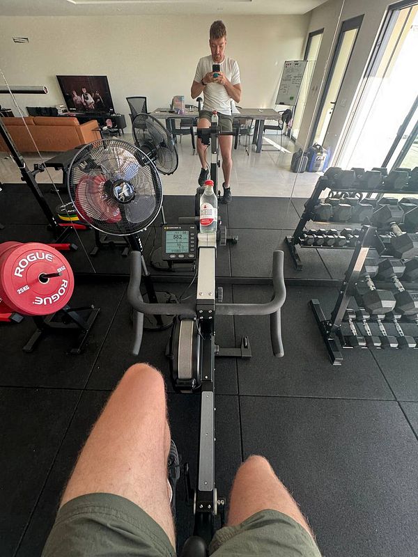 A person is seated on a rowing machine in a home gym, taking a selfie in the mirror.