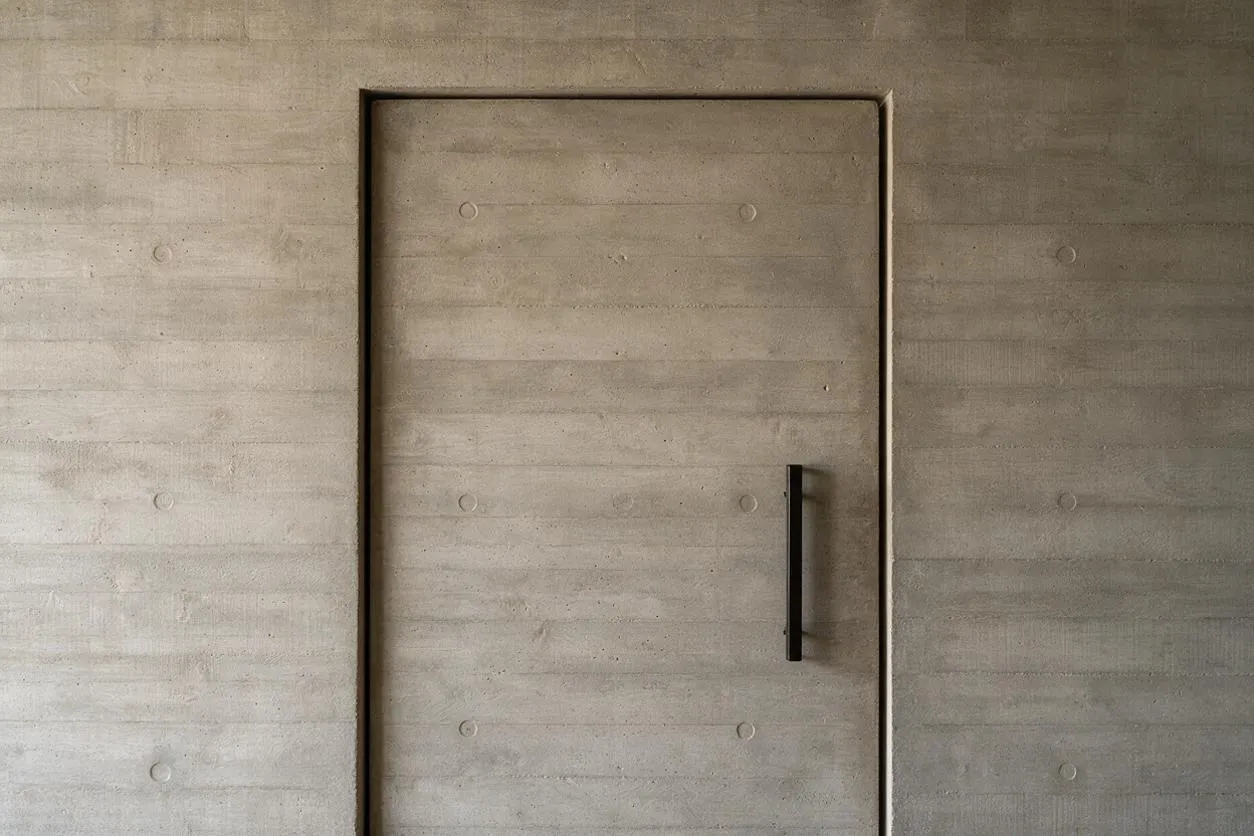 A minimalist concrete wall with a centered flush door blends into the surrounding surface. The door has a slim vertical black handle, and the exposed concrete shows horizontal formwork lines and subtle circular impressions, emphasizing the raw, sculptural Brutalist aesthetic.