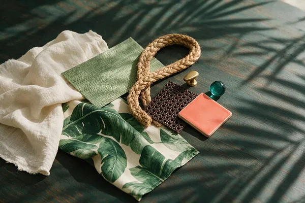 A styled flat lay of tropical-inspired materials on a dark green surface with dappled light and shadow. The arrangement includes a cream linen fabric, a green woven swatch, a palm-leaf print textile, braided rope, dark woven trim, a coral tile sample, and small brass and glass accents.